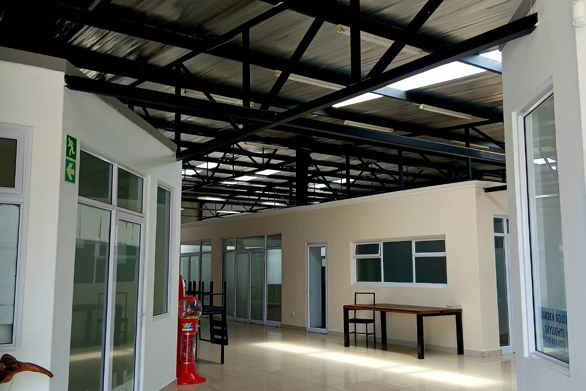 Spacious open-plan commercial interior featuring high exposed steel roof trusses, abundant natural light from skylights, and modern tiled flooring with glass partition offices.
