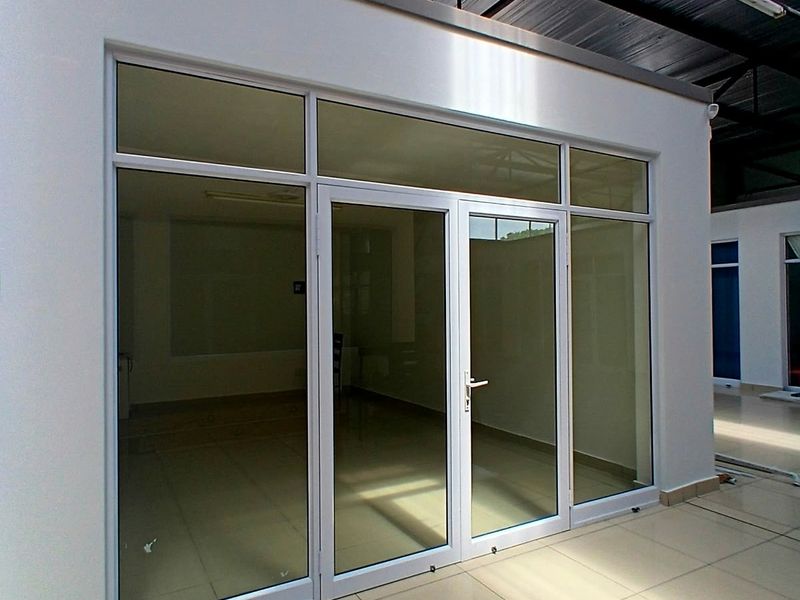 Modern office space featuring aluminium-framed glass doors, tiled flooring, and an exposed industrial-style roof structure.