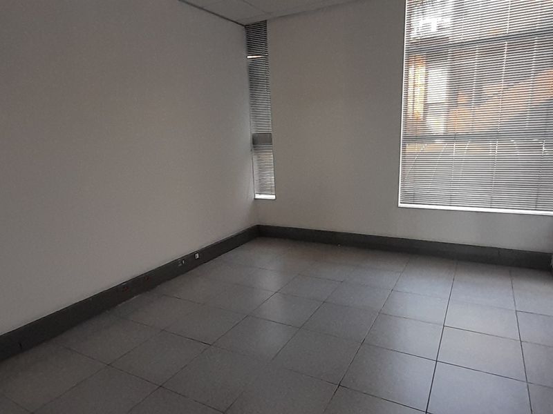 A clean, empty office space featuring tiled flooring, a suspended ceiling, and large windows fitted with venetian blinds.