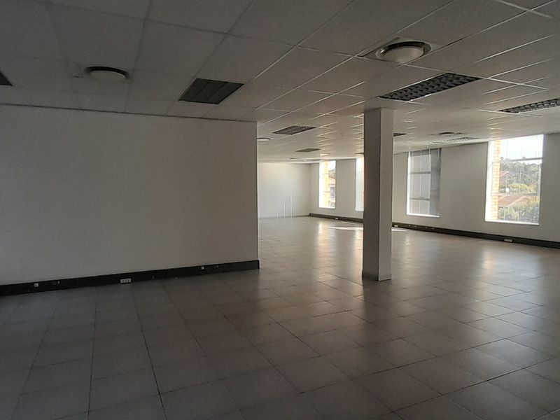 A spacious, open-plan office area featuring tiled flooring, a suspended ceiling with recessed lighting, and large windows providing ample natural light.