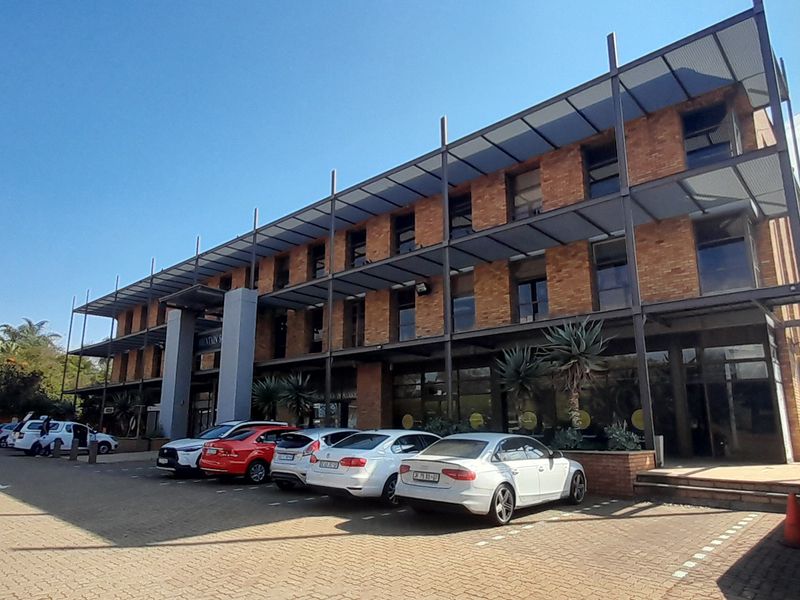 A modern multi-story office building featuring an exposed brick facade, steel structural elements, and extensive glass windows with an adjacent paved parking area.