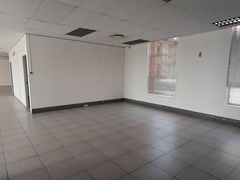 An open-plan office space featuring tiled flooring, a suspended ceiling with recessed lighting, and large windows fitted with venetian blinds.