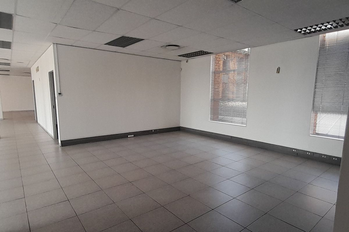 An open-plan office space featuring tiled flooring, a suspended ceiling with recessed lighting, and large windows fitted with venetian blinds.