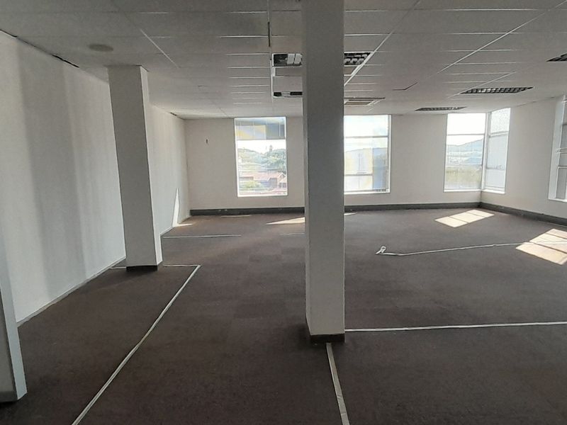 Spacious open-plan office area featuring suspended ceilings, recessed lighting, structural columns, and large windows with venetian blinds, offering a versatile workspace.