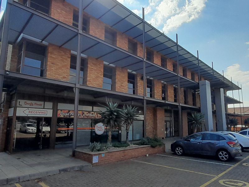 A multi-story commercial office building featuring a brick facade, steel structural framing, and a paved parking area with a covered ground-level walkway.