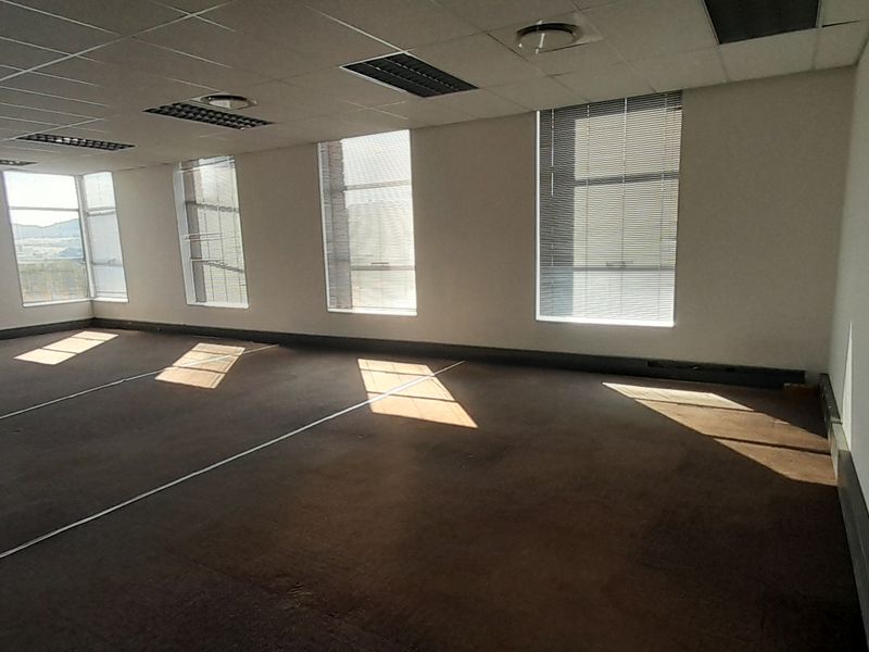 A spacious, empty open-plan office area featuring large windows with venetian blinds, recessed ceiling lighting, and carpeted flooring.
