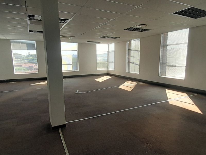 A spacious, open-plan office area featuring large windows with blinds, carpeted flooring, and a suspended ceiling with recessed lighting.