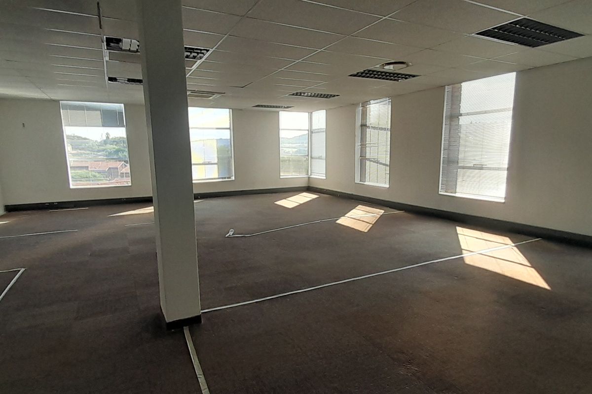 A spacious, open-plan office area featuring large windows with blinds, carpeted flooring, and a suspended ceiling with recessed lighting.