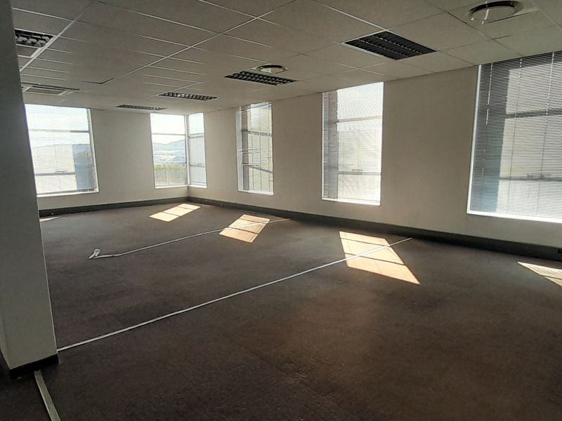 A spacious, empty open-plan office area featuring large windows with blinds, suspended ceilings, and carpeted flooring, offering a versatile workspace.