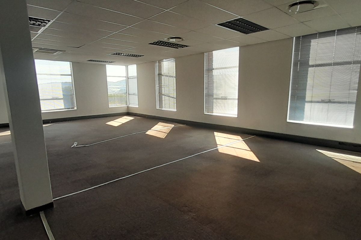 A spacious, empty open-plan office area featuring large windows with blinds, suspended ceilings, and carpeted flooring, offering a versatile workspace.