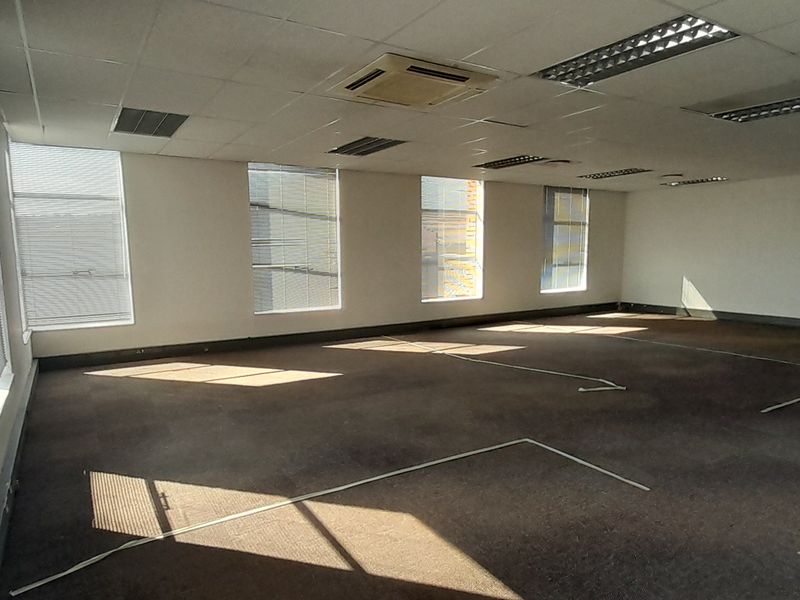 Spacious open-plan office area featuring suspended ceilings, recessed lighting, central air conditioning, and large windows with venetian blinds.