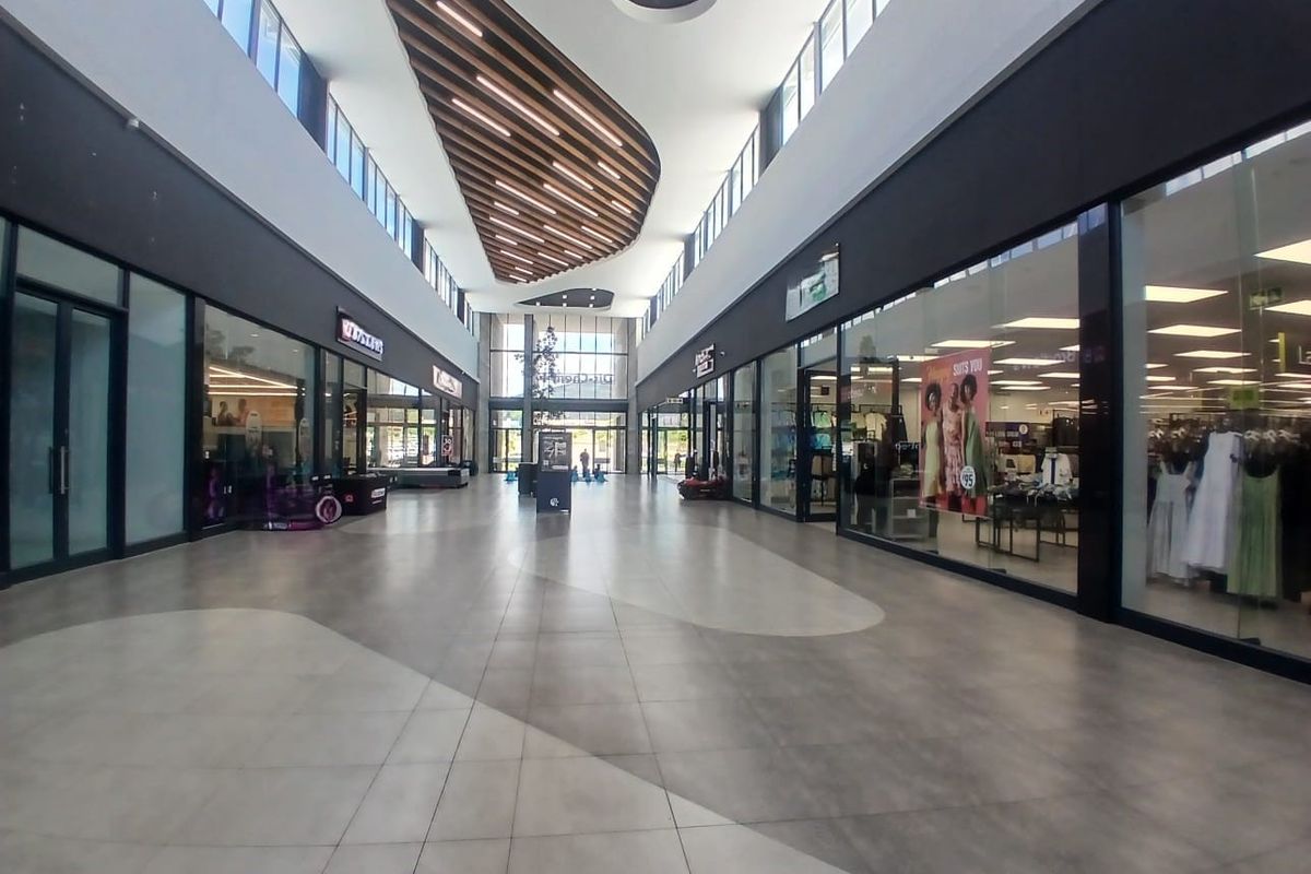 Modern retail shopping centre corridor featuring high ceilings with decorative timber accents, large glass storefronts, and polished tiled flooring.