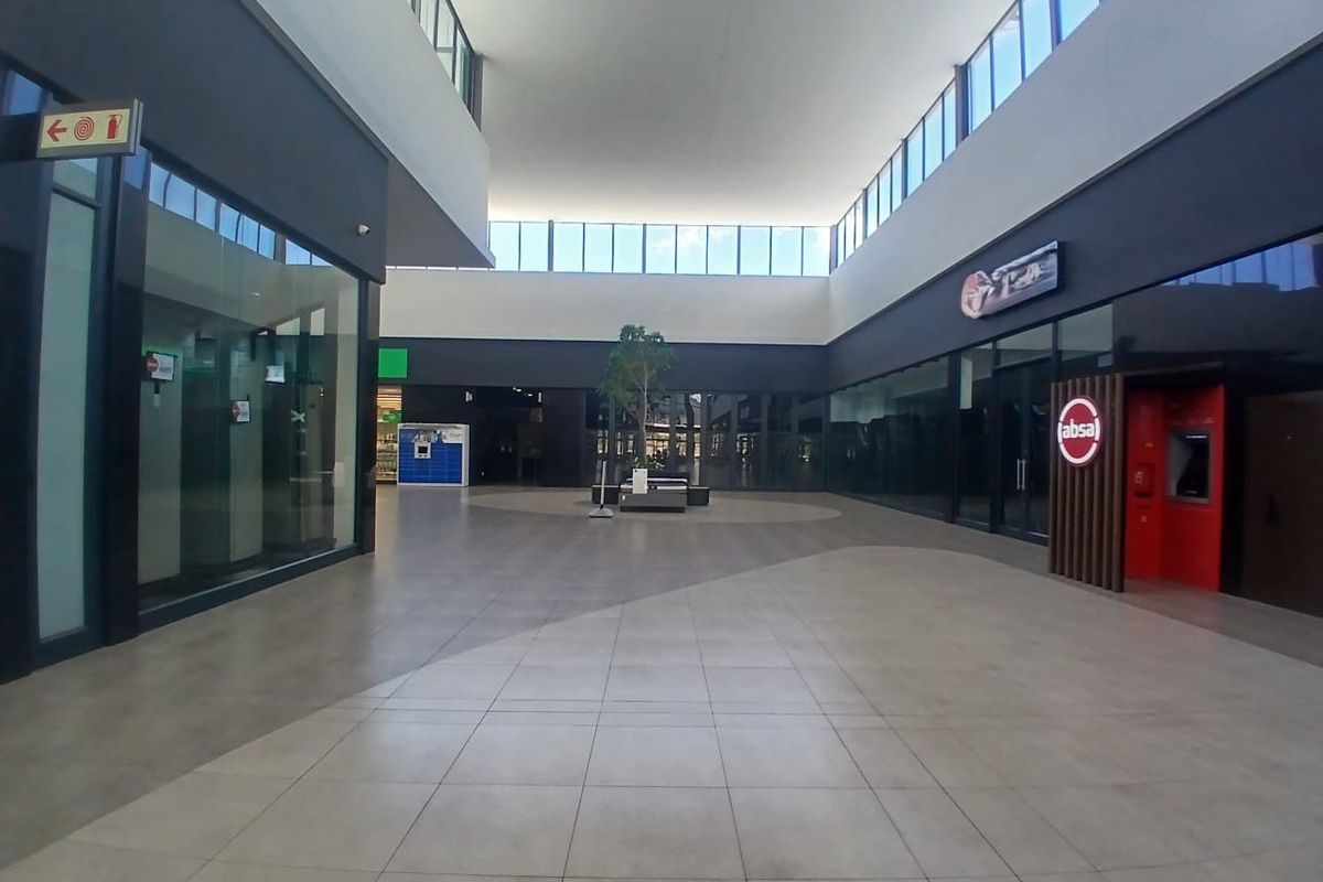 A spacious, modern retail mall corridor featuring large glass storefronts, high ceilings with clerestory windows, and clean tiled flooring.