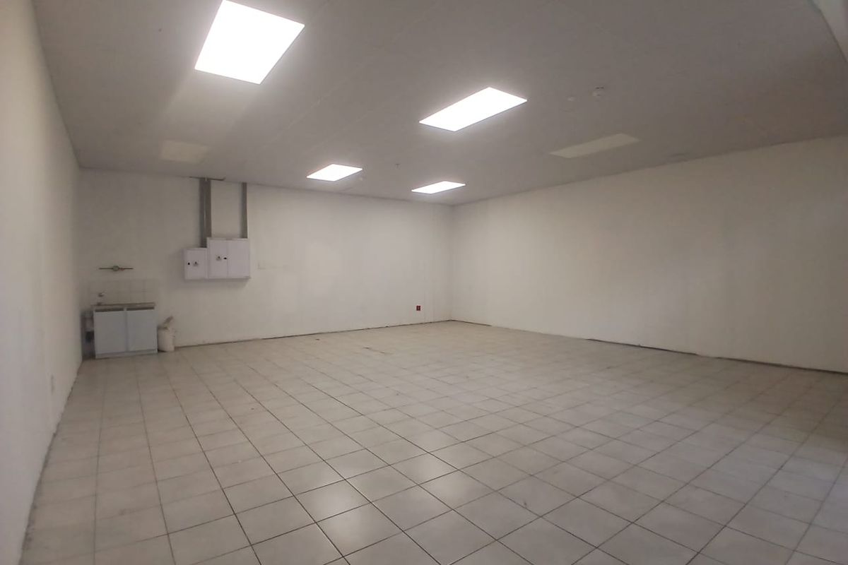 A spacious, empty open-plan retail unit featuring tiled flooring, recessed ceiling lighting, and a small kitchenette area, suitable for various commercial uses.