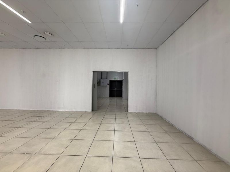 A vacant, open-plan retail space featuring neutral ceramic floor tiles, a suspended ceiling with integrated lighting, and a rear fire exit door.