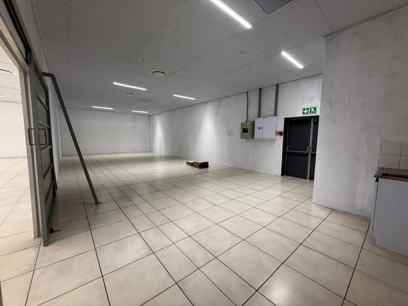A spacious, empty open-plan retail unit featuring tiled flooring, suspended ceilings with recessed lighting, and a small kitchenette area.