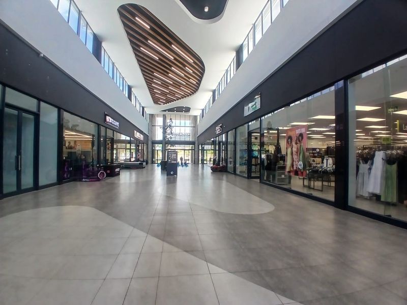 Modern retail shopping centre corridor featuring high ceilings with decorative timber accents, large glass storefronts, and polished tiled flooring.