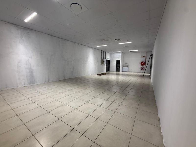 An empty, open-plan retail utility space featuring tiled flooring, a suspended ceiling with recessed lighting, and basic kitchenette facilities.