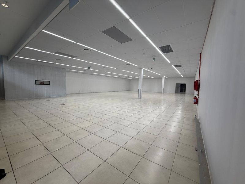 A spacious, vacant open-plan retail unit featuring neutral ceramic floor tiles, a suspended ceiling with integrated linear lighting, and essential fire safety equipment.