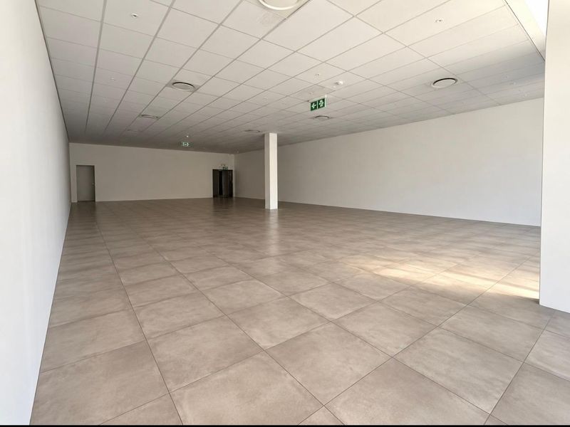 A spacious, vacant open-plan retail unit featuring neutral tiled flooring, a suspended ceiling with recessed lighting, and a central structural column.