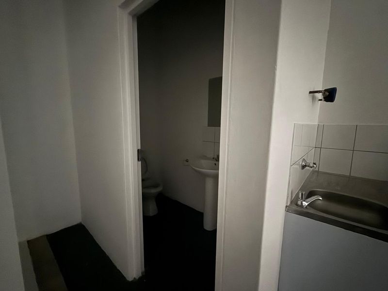 A small, dimly lit bathroom featuring a pedestal sink and toilet, with an adjacent utility area containing a stainless steel basin.
