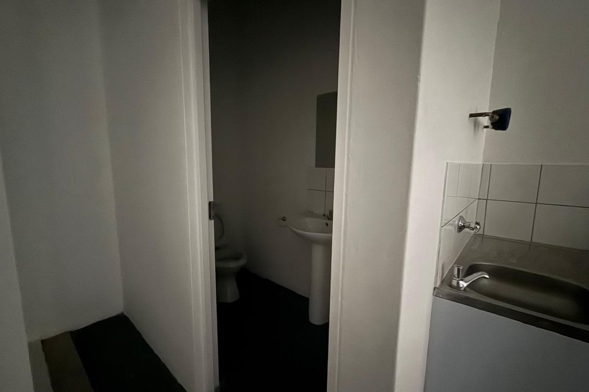 A small, dimly lit bathroom featuring a pedestal sink and toilet, with an adjacent utility area containing a stainless steel basin.