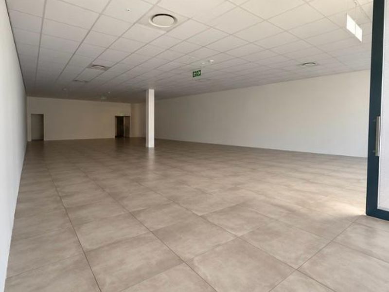A spacious, vacant open-plan retail unit featuring neutral tiled flooring, a clean suspended ceiling with recessed lighting, and a central support column.