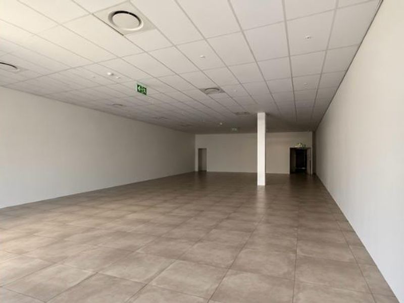 A spacious, vacant retail unit featuring neutral ceramic floor tiles, a clean suspended ceiling with recessed lighting, and an open-plan layout.
