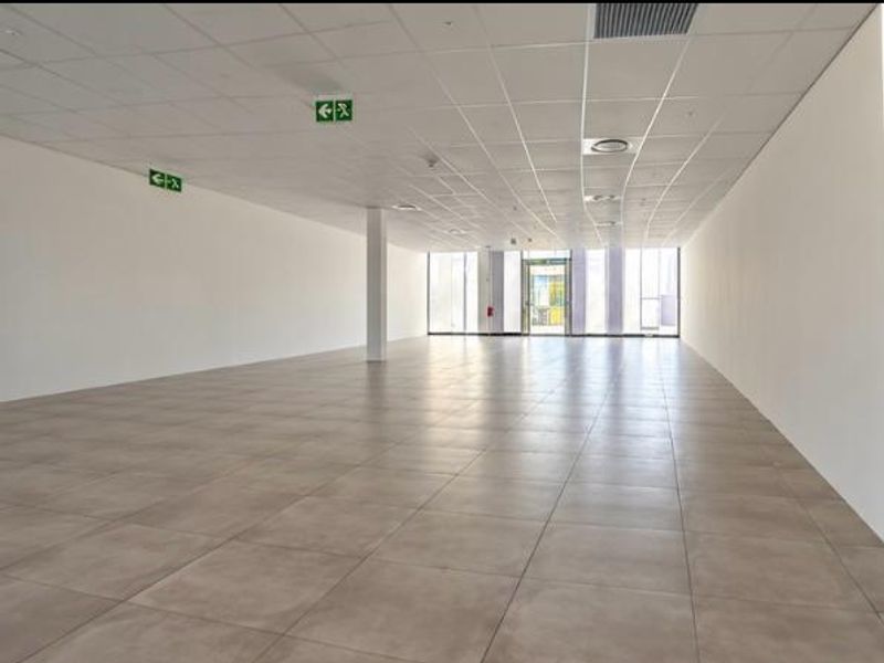 A spacious, open-plan retail unit featuring neutral tiled flooring, a clean suspended ceiling with recessed lighting, and large glass windows providing ample natural light.