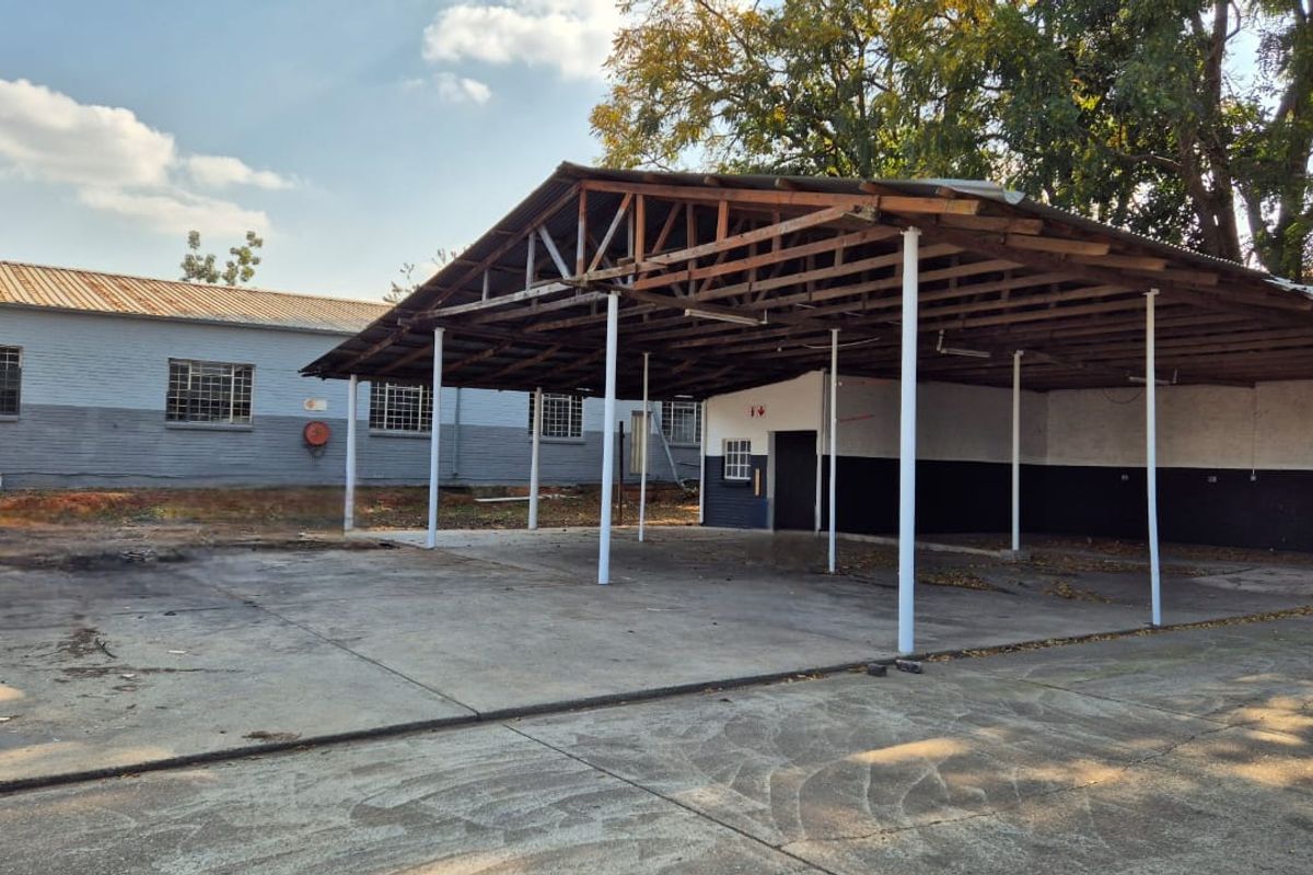 Industrial exterior area featuring a large concrete hardstand and a covered steel-framed canopy structure with security-barred windows on the adjacent building.