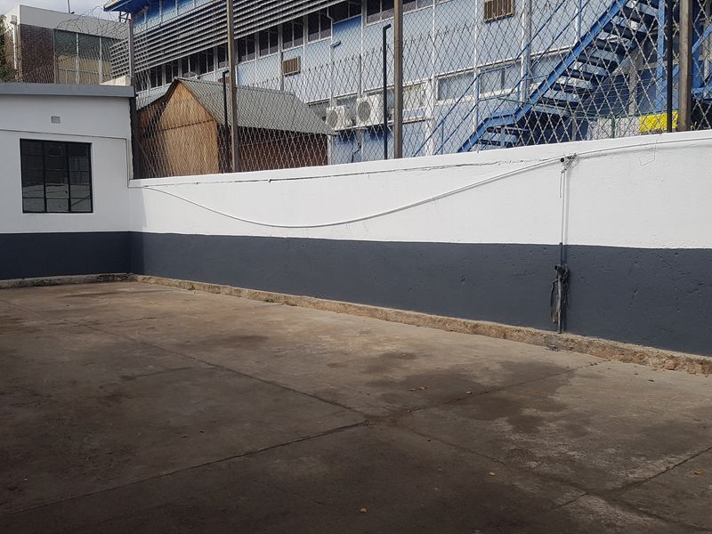 A paved industrial rear yard area featuring a perimeter wall with security fencing and a small utility structure.