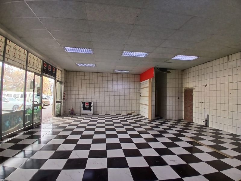 An empty retail space featuring a checkered tiled floor, suspended ceiling with recessed lighting, and large glass storefront windows.