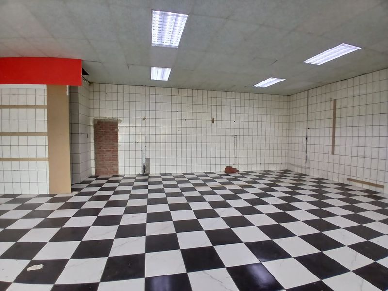 An empty retail space featuring a black and white checkered tiled floor, tiled walls, and recessed fluorescent lighting, requiring cosmetic refurbishment.