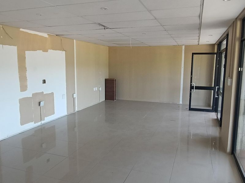 An empty open-plan retail space featuring tiled flooring, a suspended ceiling, and large glass doors leading to an outdoor area.