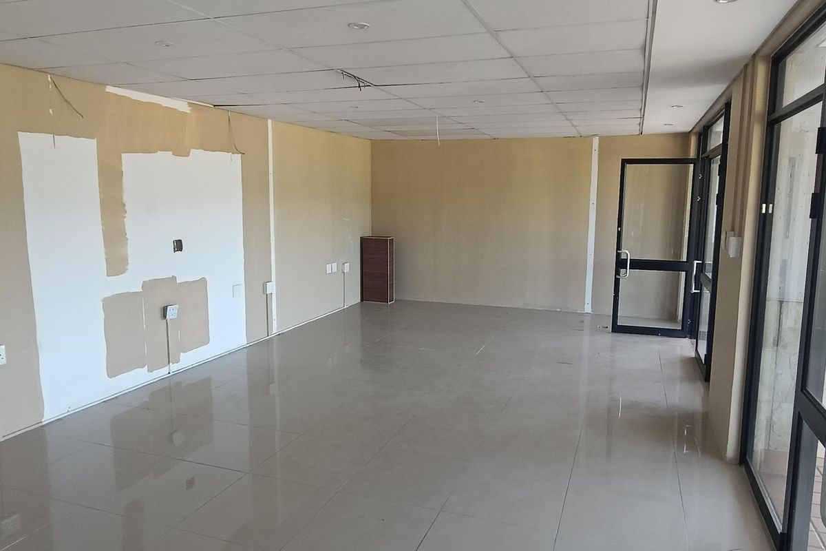 An empty open-plan retail space featuring tiled flooring, a suspended ceiling, and large glass doors leading to an outdoor area.