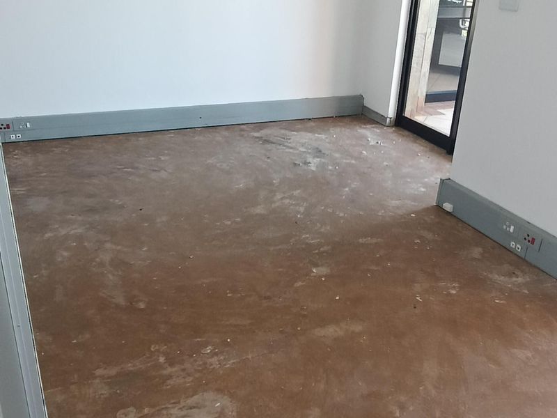 An empty office space featuring a suspended ceiling, recessed lighting, and perimeter trunking, currently awaiting final flooring installation.