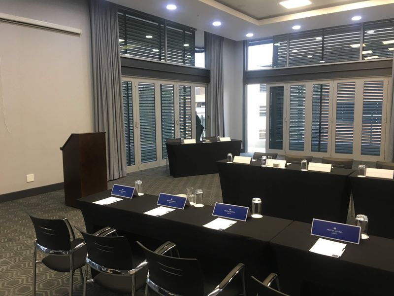 A professional office meeting room featuring recessed lighting, large windows with louvred shutters, and carpeted flooring, set up for a corporate conference.