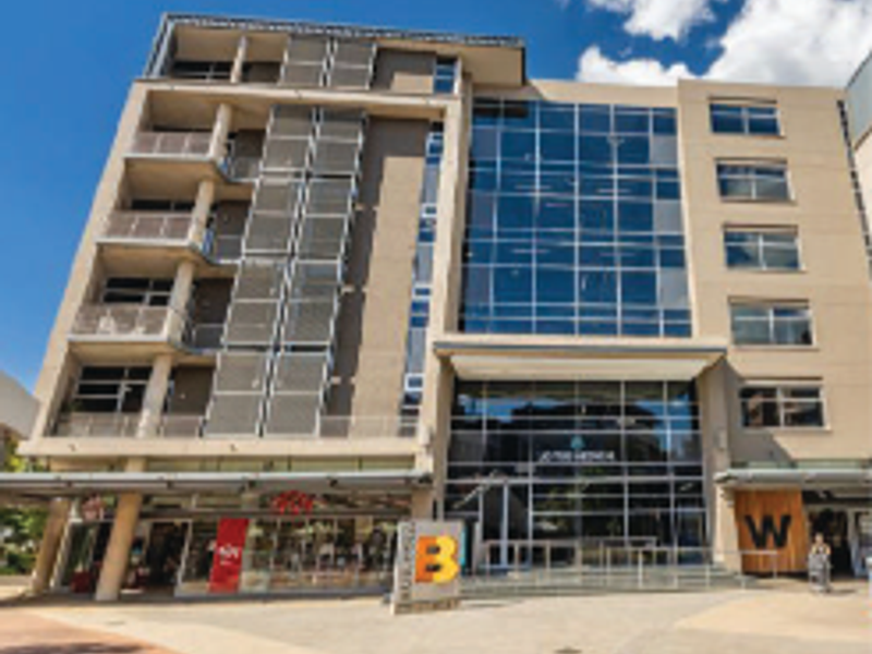 Modern multi-story office building featuring a glass facade, integrated balconies with metal louvers, and ground-level commercial retail spaces.