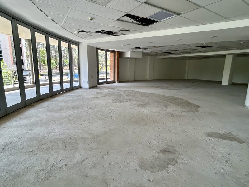 Spacious open-plan office area featuring large glass doors, structural columns, and a suspended ceiling requiring repair and flooring installation.