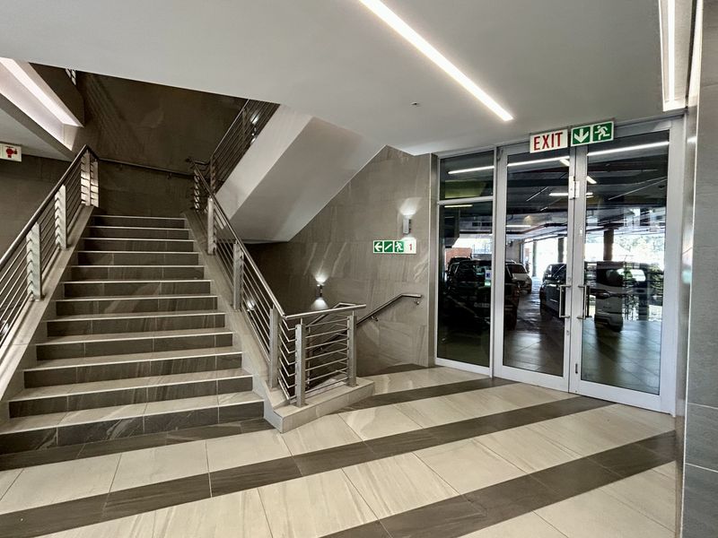 Modern office building hallway featuring a tiled staircase with stainless steel handrails and glass double doors leading to the parking area.