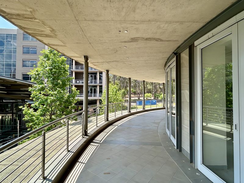 A spacious, curved balcony featuring tiled flooring, a concrete ceiling with recessed lighting, and metal railings, offering views of the surrounding office complex.