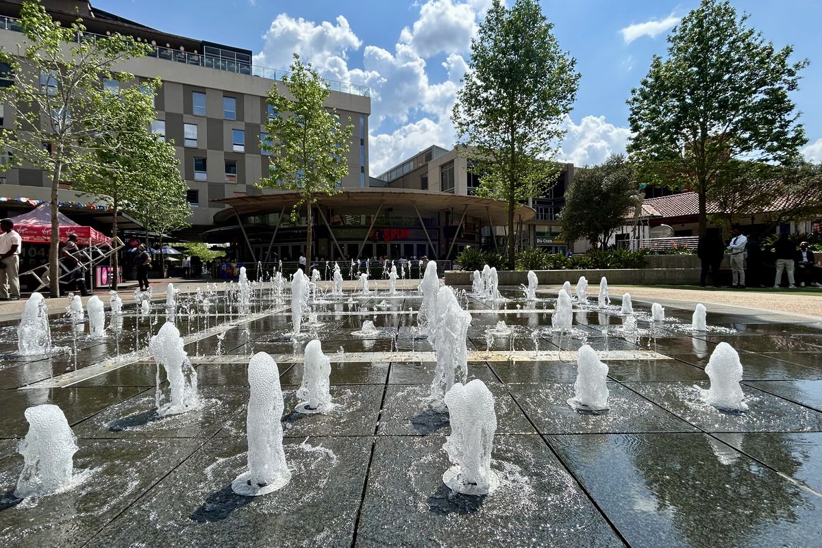 A vibrant outdoor plaza featuring a modern water fountain, paved walkways, and surrounding commercial office buildings in a well-maintained landscaped environment.