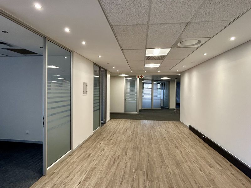 A bright, modern office hallway featuring wood-look flooring, recessed lighting, and glass partition walls leading to multiple private office spaces.