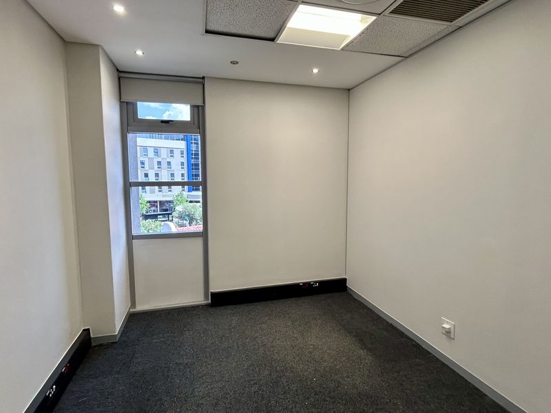 A clean, empty office space featuring neutral walls, carpet flooring, recessed lighting, and a window with a roller blind.