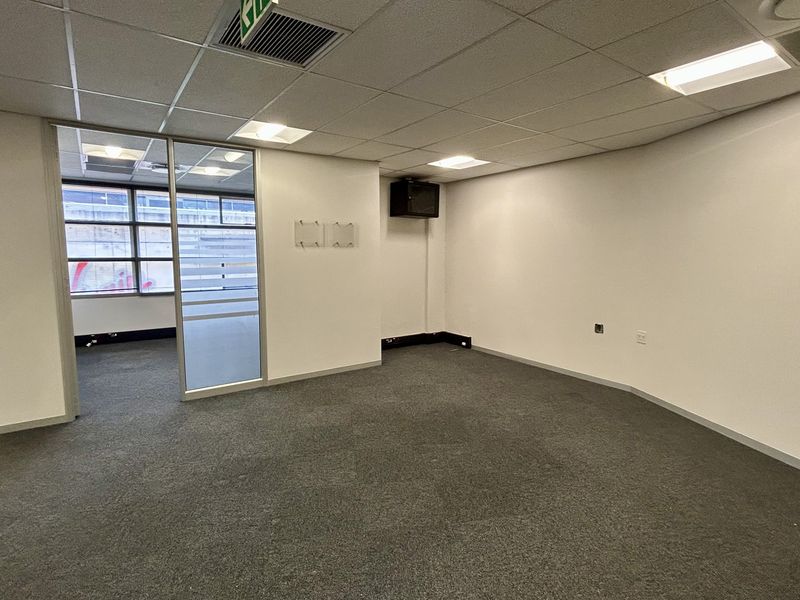An empty, open-plan office space featuring neutral carpet flooring, a suspended ceiling with recessed lighting, and a glass partition wall.