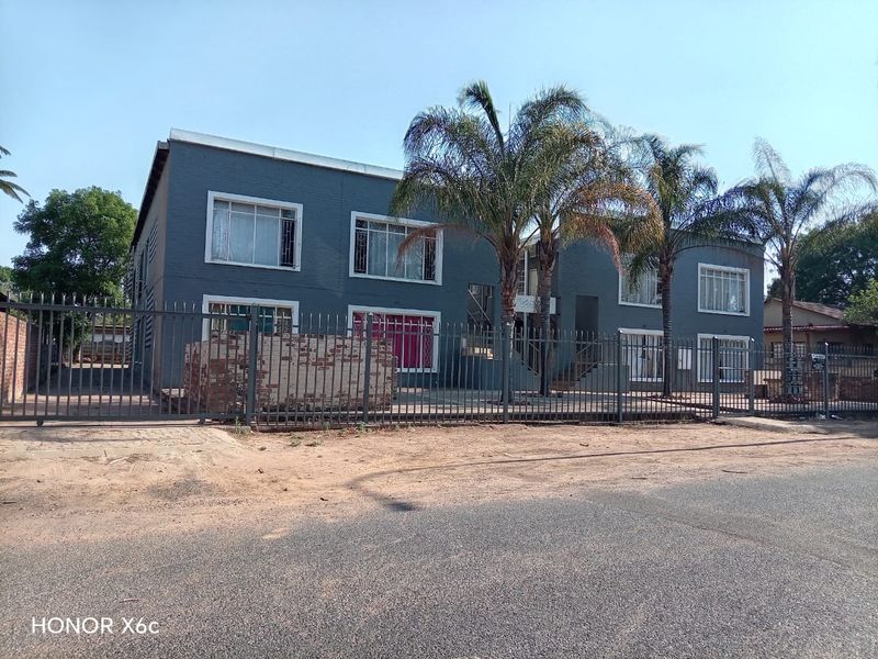 Street-facing view of a two-story apartment complex featuring dark grey painted brick facade, metal security fencing, and established palm trees.
