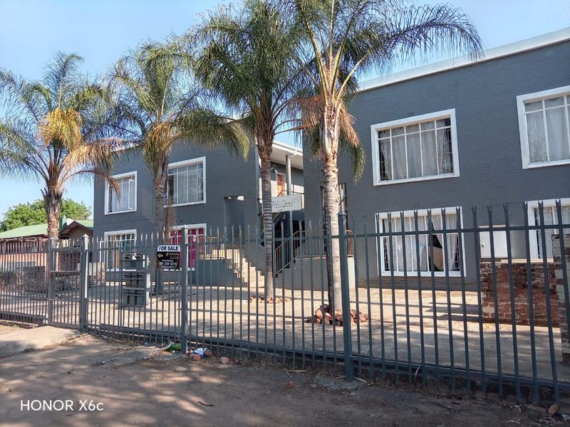 Exterior view of a multi-story apartment complex featuring grey painted brick facade, security fencing, and palm trees in the front courtyard area.