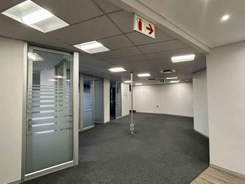 A spacious, vacant open-plan office area featuring modern glass partitions, suspended ceilings with recessed lighting, and neutral carpet flooring.