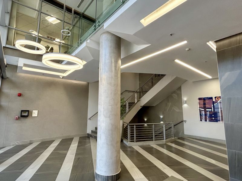 Modern office building entrance lobby featuring a double-volume ceiling, glass balustrades, a central concrete column, and high-quality tiled flooring.