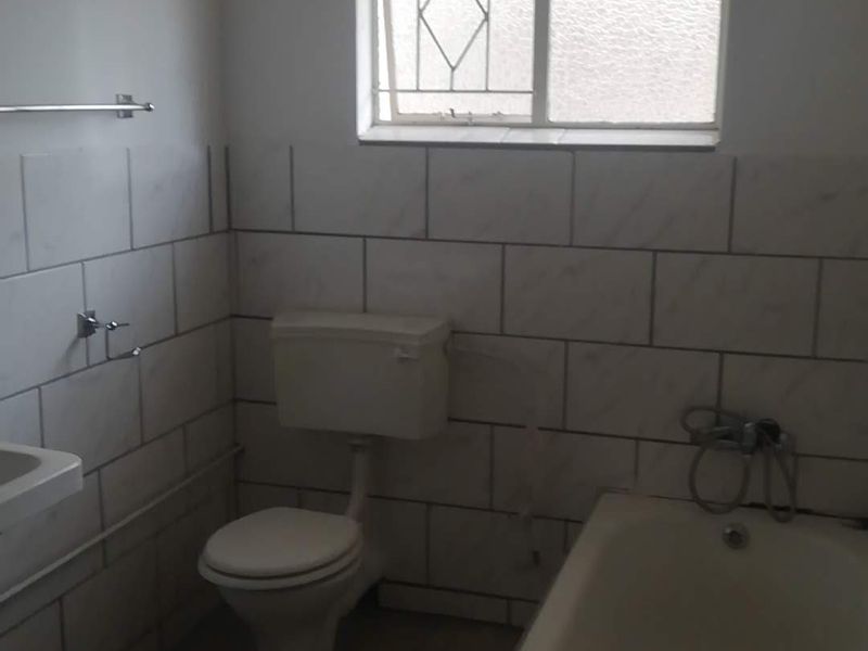 A functional bathroom featuring a bathtub, toilet, and wall-mounted basin with tiled walls and a window with security bars.